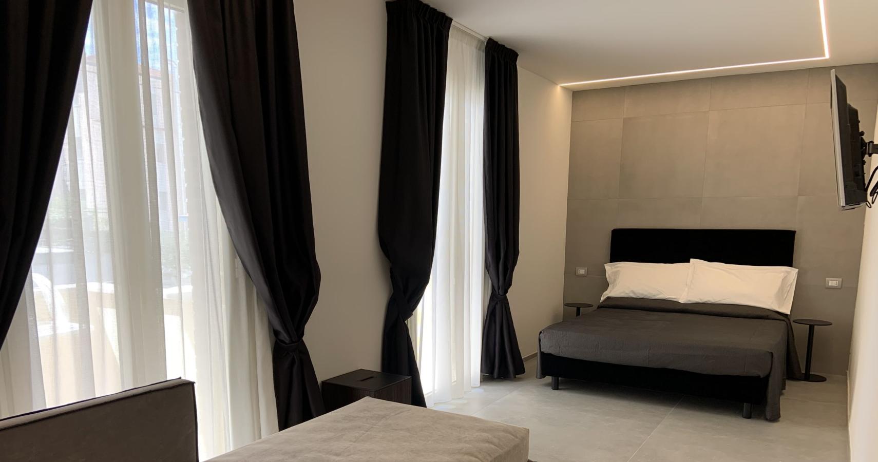 Camera moderna con letto matrimoniale, divano, tende nere e illuminazione soffusa.