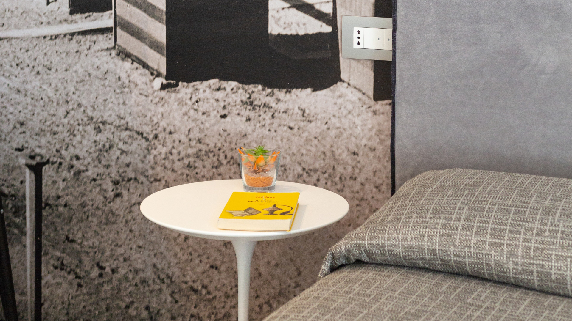 Tavolino bianco con libro giallo e pianta, accanto a un letto grigio.