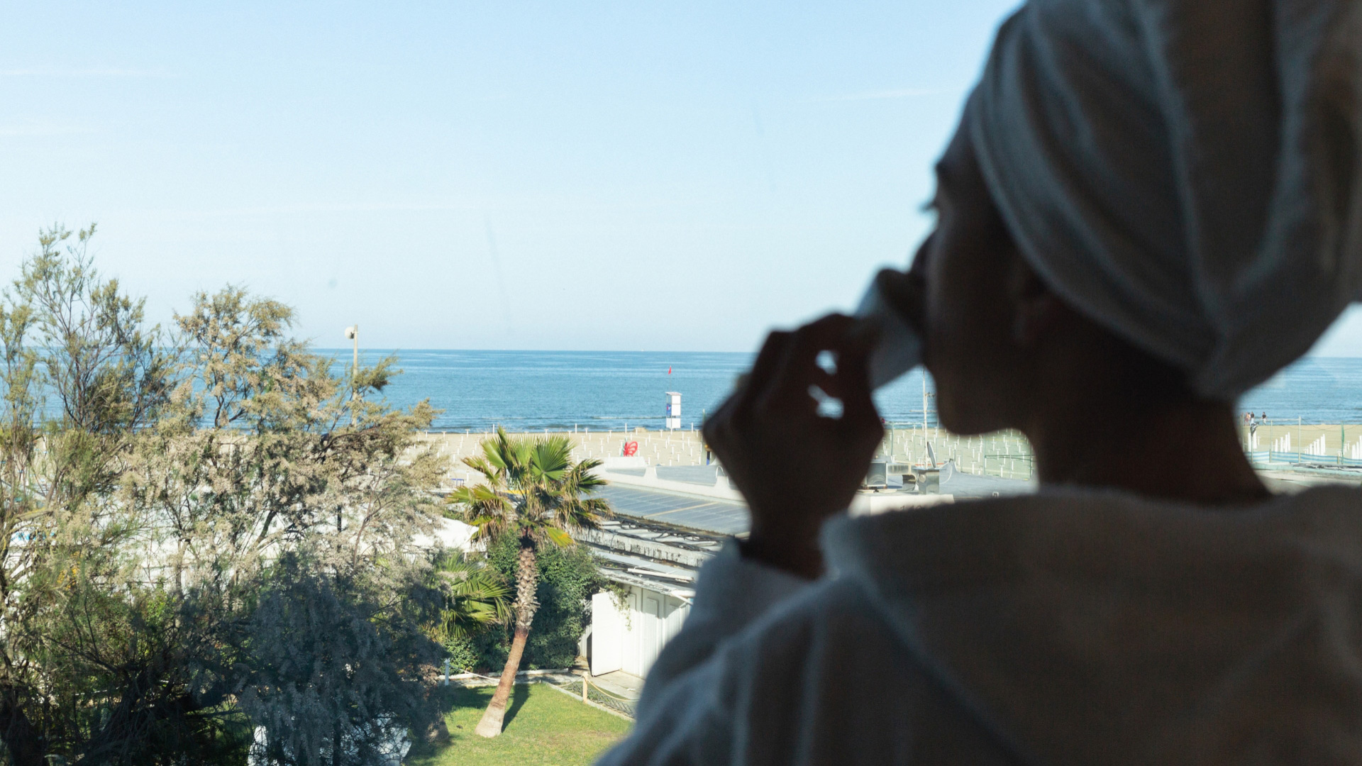 Persona in accappatoio guarda il mare da una finestra, con tazza in mano.
