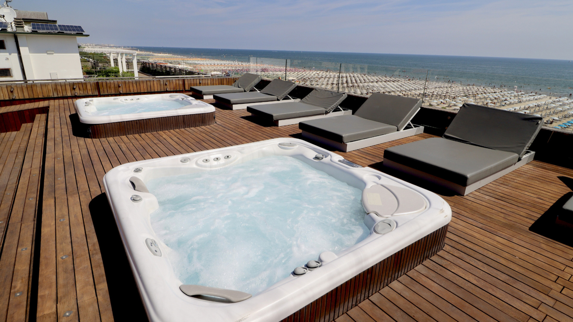 Terrazza con vasche idromassaggio e lettini, vista mare e spiaggia affollata.