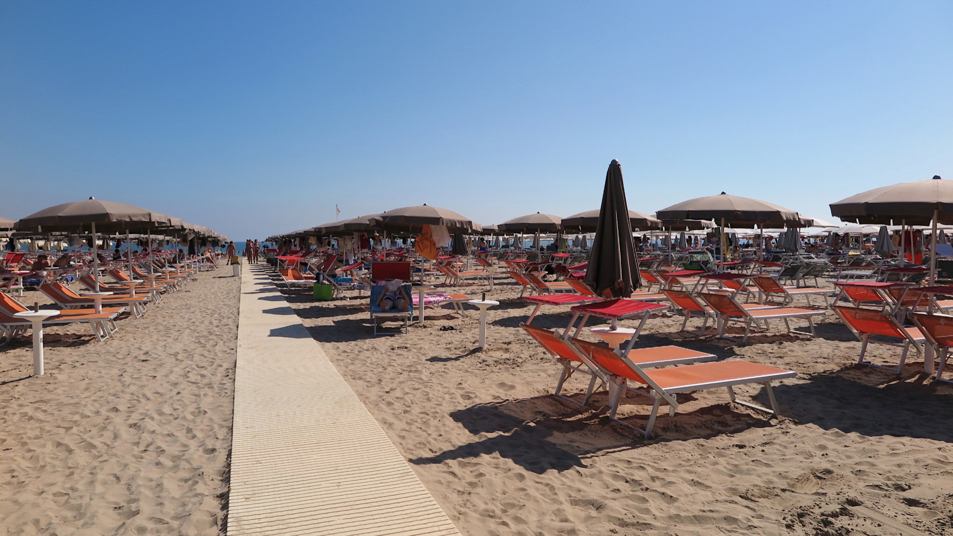 Spiaggia con ombrelloni e lettini arancioni, cielo sereno e sabbia chiara.