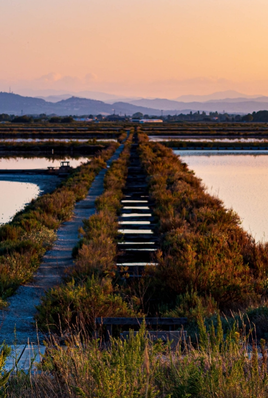 Tramonto su saline con passerella e montagne sullo sfondo.