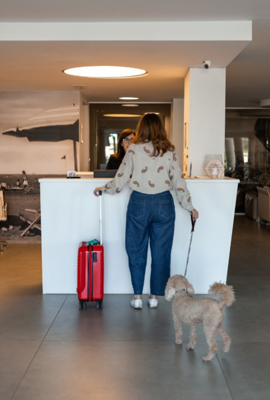 Donna con cane e valigia rossa al banco della reception di un hotel.