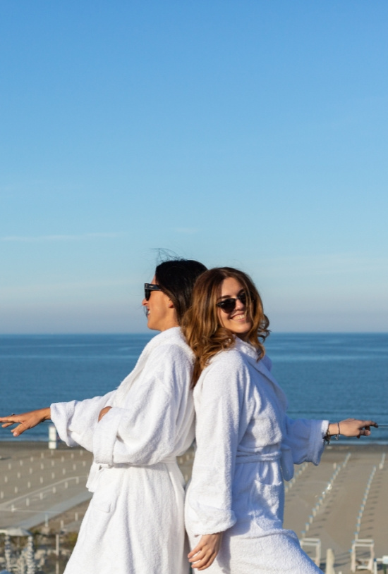 Due persone in accappatoi bianchi sorridono in spiaggia con il mare sullo sfondo.