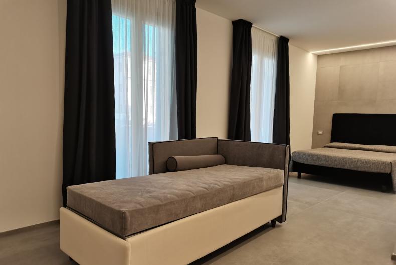 Camera moderna con divano letto e letto matrimoniale, tende scure e pavimento in cemento.