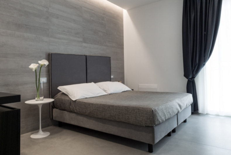 Camera minimalista con letto grigio, pareti moderne e fiori su tavolino bianco.