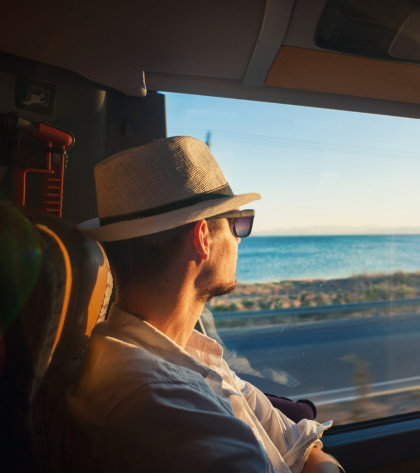 Uomo con cappello guarda il mare dal finestrino di un autobus al tramonto.