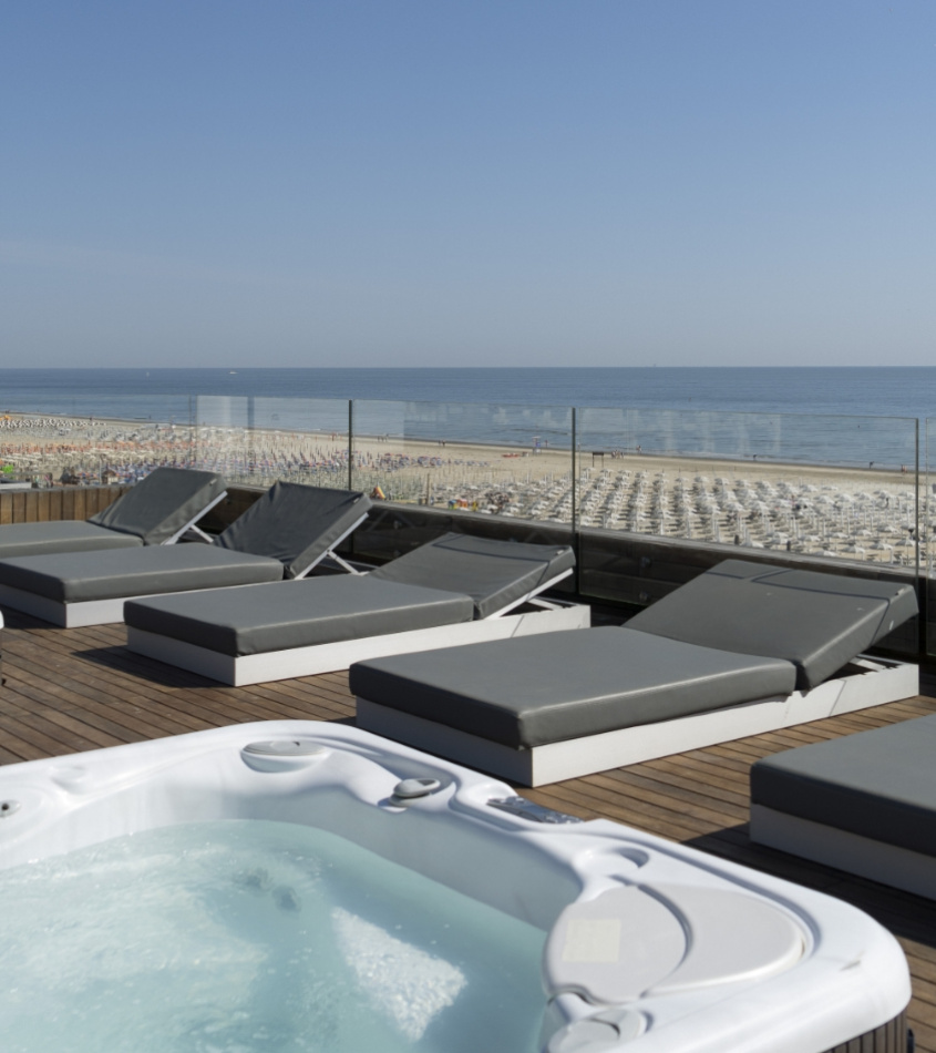 Terrazza con vasca idromassaggio, lettini e vista mare su una spiaggia affollata.